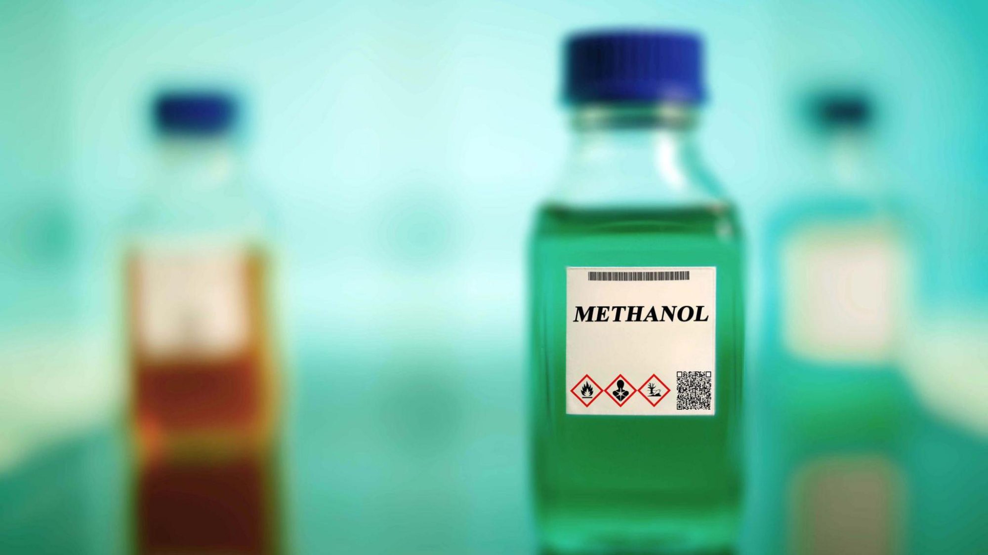 Três garrafas de vidro com uma central rotulada METANOL e pictogramas GHS de periculosidade, ilustrando o risco da adulteração de bebidas